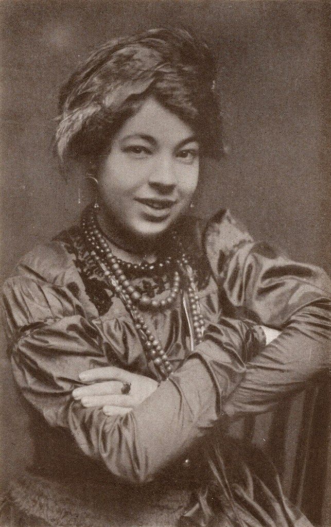 Retrato en blanco y negro de Pamela Colman Smith, una mujer con un peinado elaborado, vestida con ropa de época y con múltiples collares, sonriendo mientras cruza los brazos.